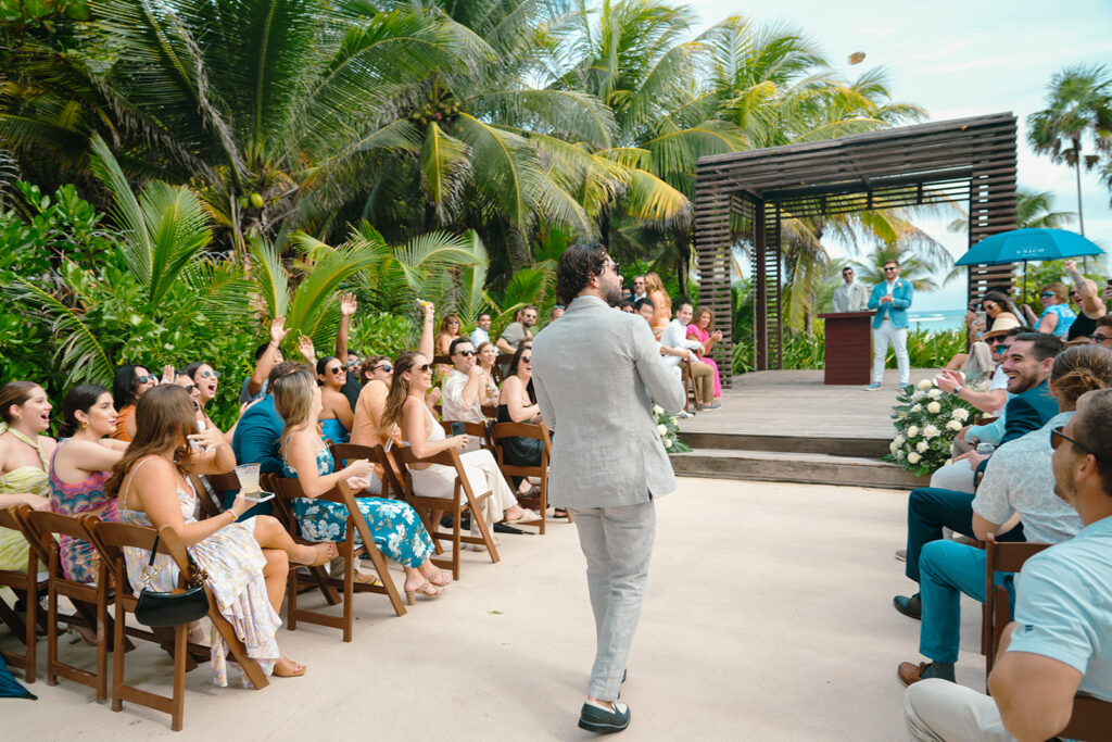 Best Man walking down aisle tossing out mini liquor bottles during wedding ceremony at UNICO Hotel Riviera Maya