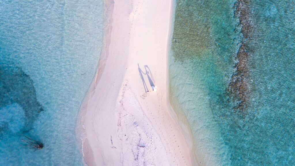 Couple in wedding attire walking down a sand bar in the ocean, viewed from above. 