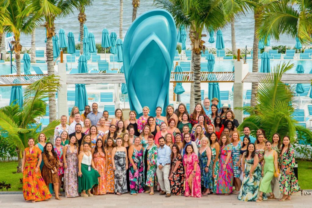 group of people wearing bright colors and patterns posing in front of flip flop statue at tropical resort